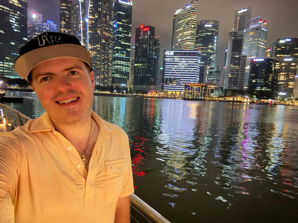 Selfie along the Singapore River at night, CBD lights behind, Rays cap.